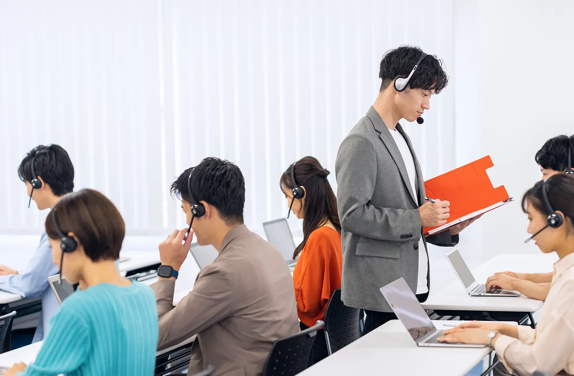 ヘッドセットを付けたスタッフを見守りながら、資料を確認するリーダーと業務中のチーム