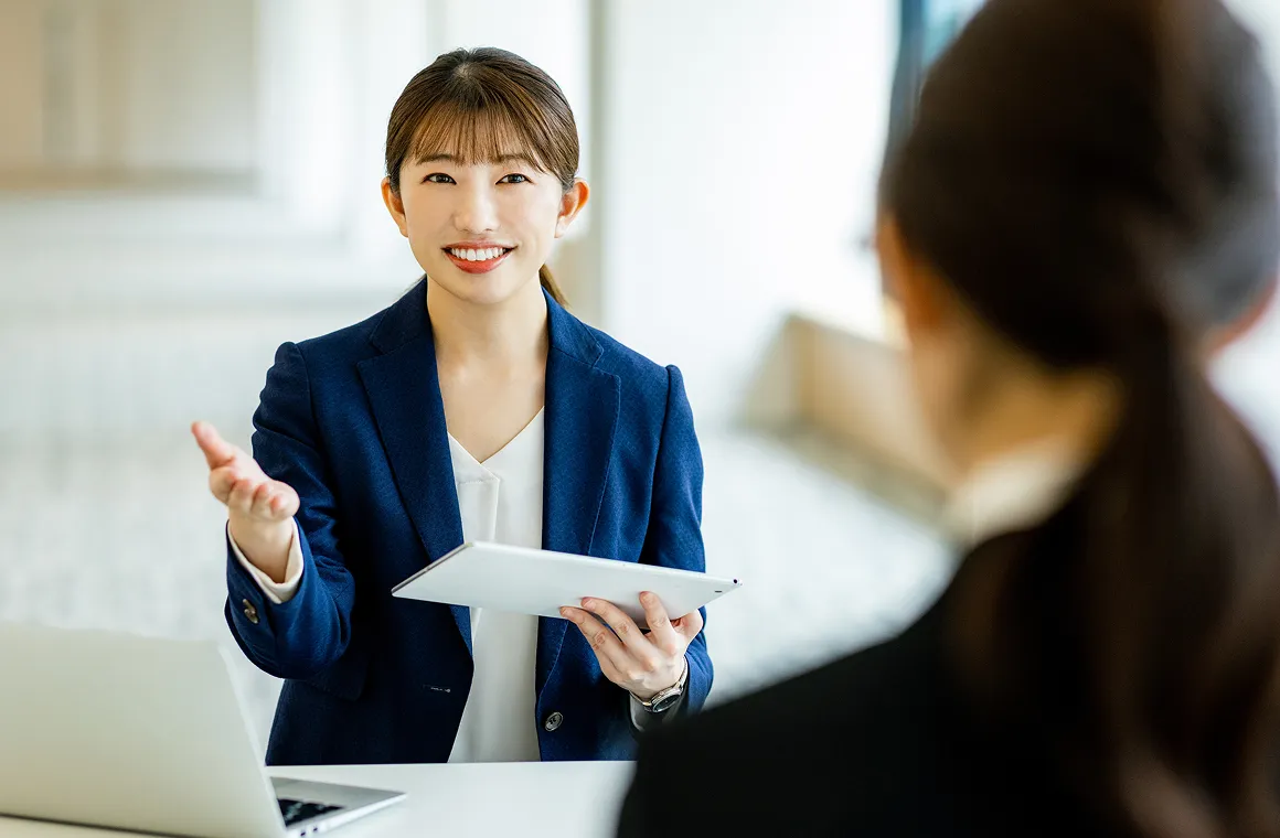 タブレットを手に、相手に向かって説明を行うスーツ姿の女性スタッフ