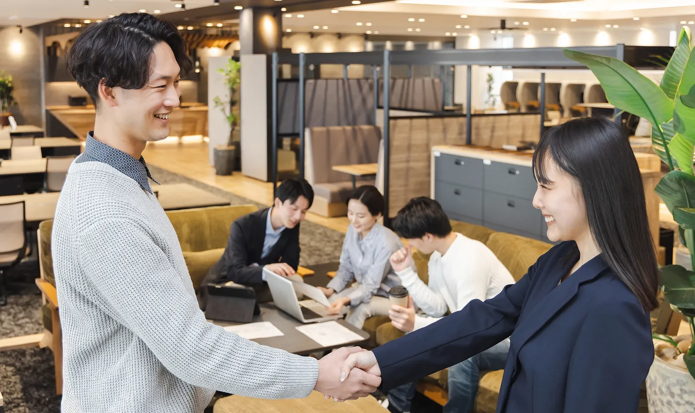 オフィスの共有スペースで、笑顔で握手を交わす男女と、後ろで打ち合わせを行うチームメンバー