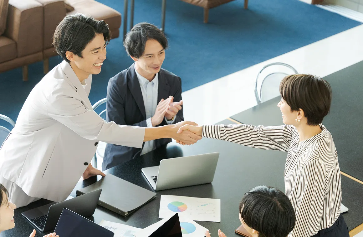 会議テーブル越しに握手をする二人と拍手する同席者のいるミーティング風景