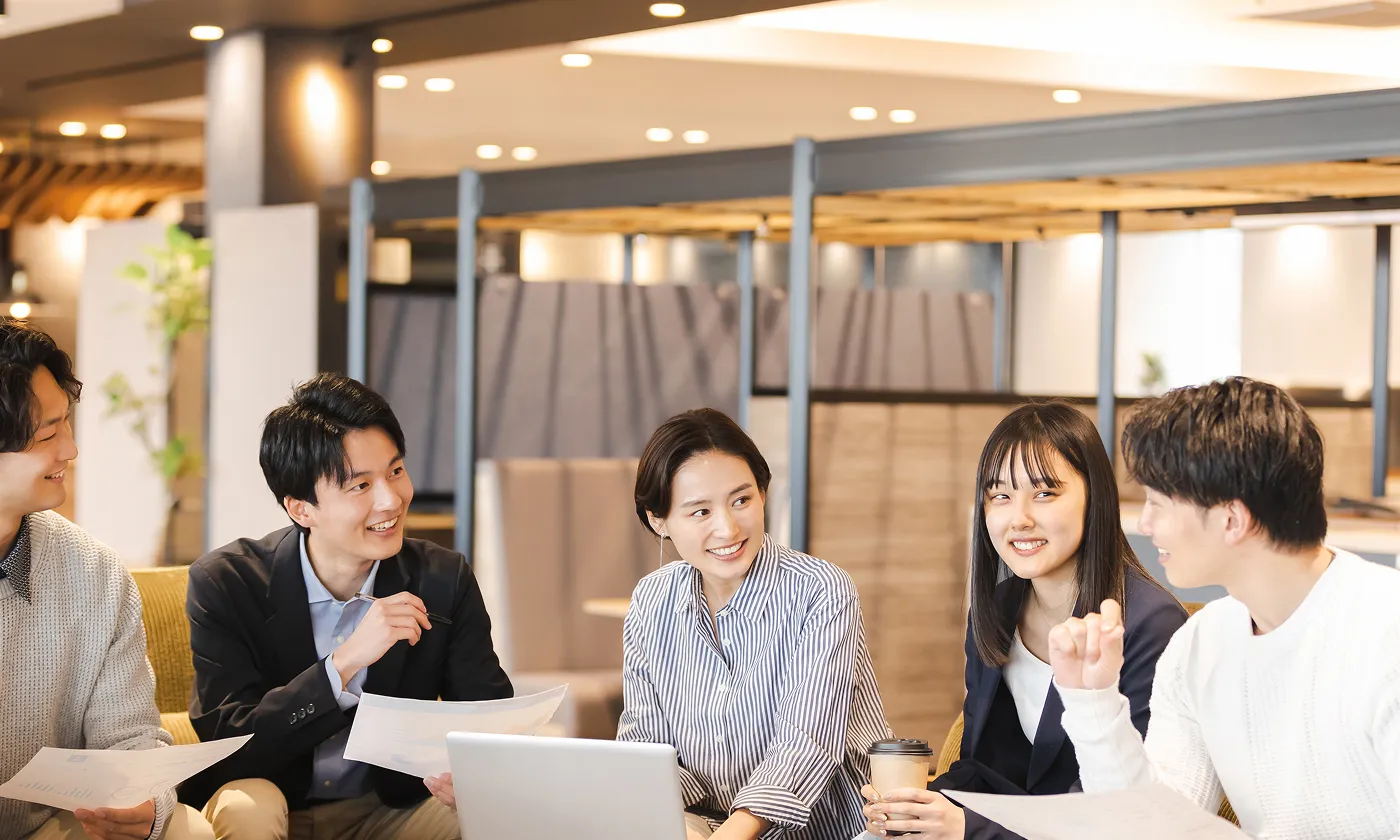 オフィスの共有スペースで、資料とノートパソコンを囲みながら意見交換する若手メンバーの打ち合わせ風景