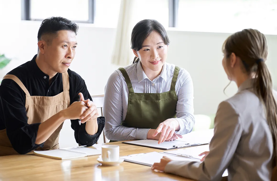 テーブルを囲み、書類を見ながら相談を受けるエプロン姿の男女とスーツ姿の女性