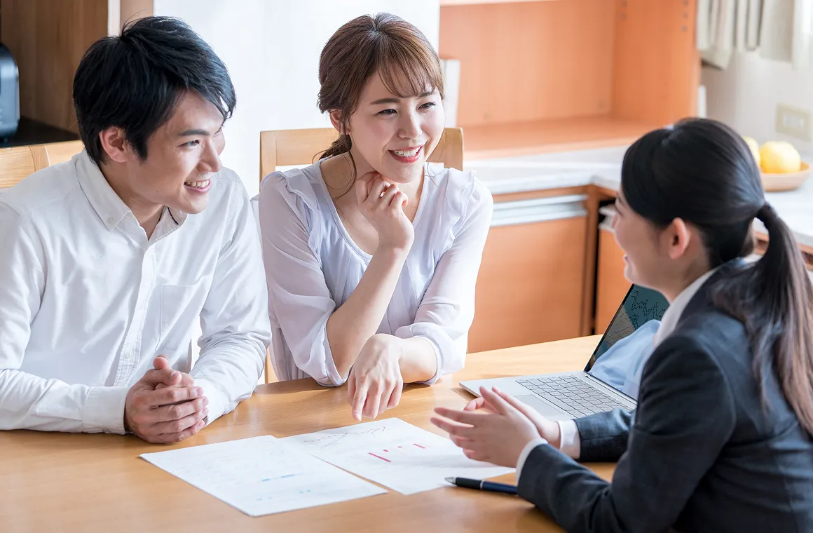資料を広げたテーブルで、笑顔で説明を受ける男女とノートパソコンを使う担当者