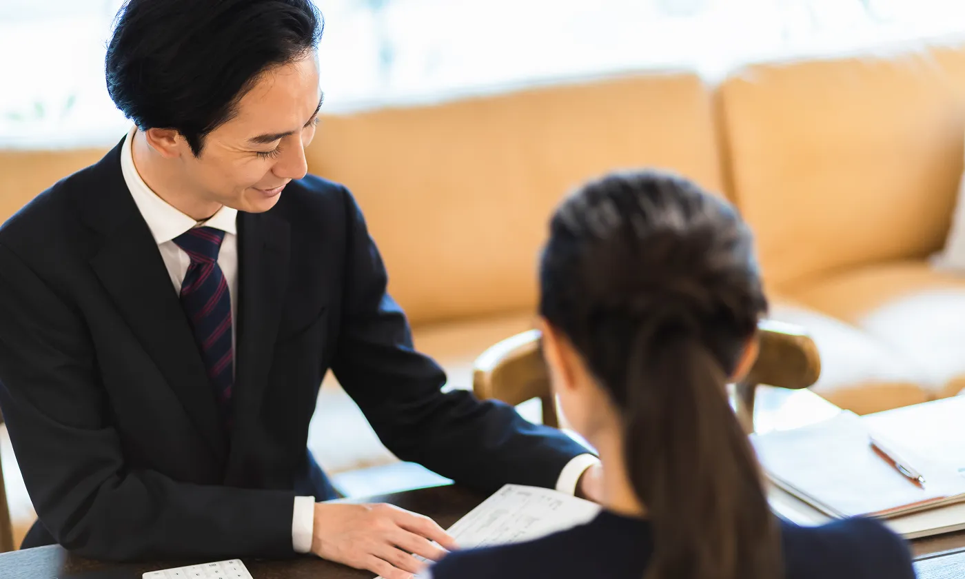 ソファのある空間で、書類を指し示しながら丁寧に説明するスーツ姿の男性