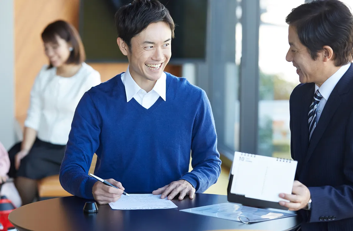テーブルで書類に記入しながら、スーツ姿の男性と談笑する男性