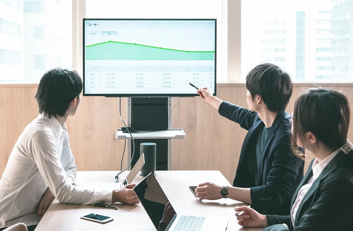 モニターのグラフを指し示しながらデータ分析について議論するミーティング風景