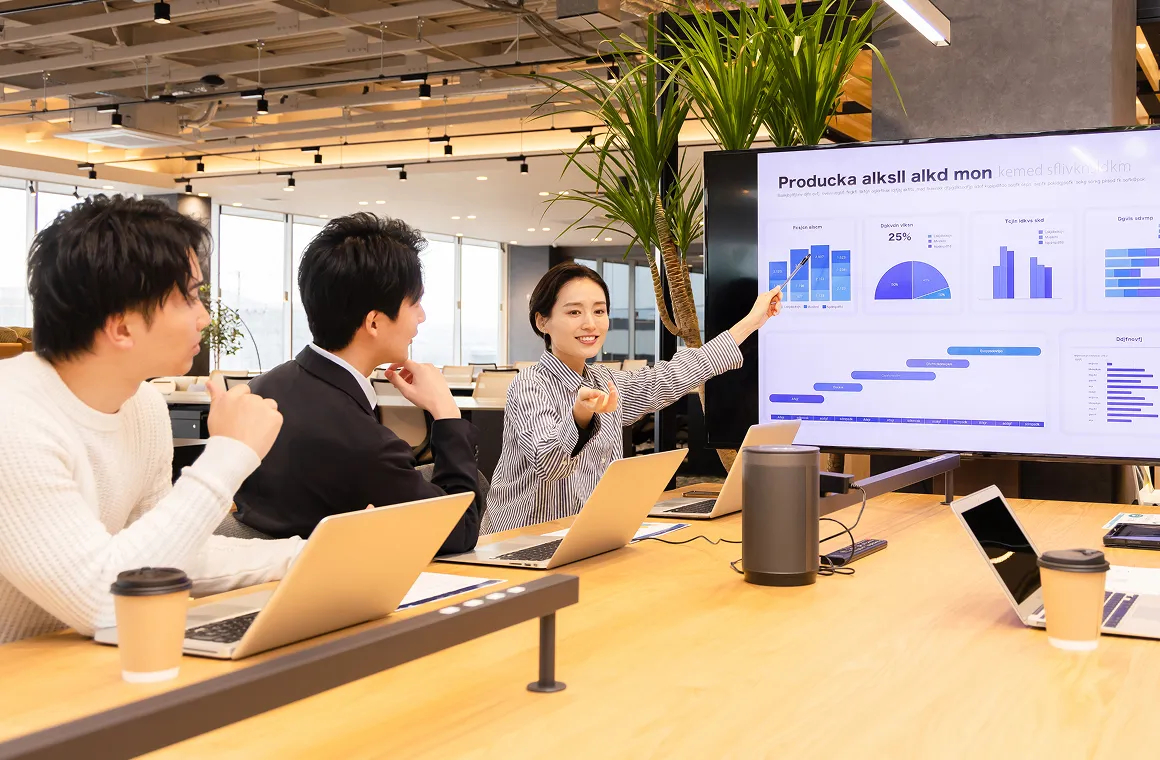 大型モニターのグラフを指し示しながらプレゼンテーションを行う女性と打ち合わせ中のチーム