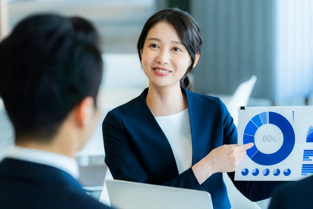 スーツ姿の二人がテーブルで資料やノートパソコンを前に商談や打ち合わせをしている様子